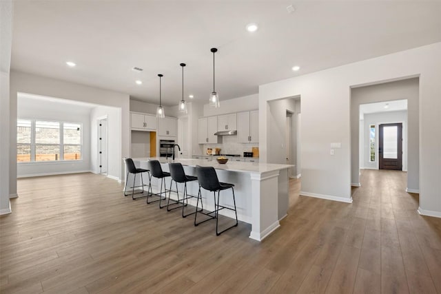 kitchen with light wood-style flooring, under cabinet range hood, white cabinets, light countertops, and a large island with sink