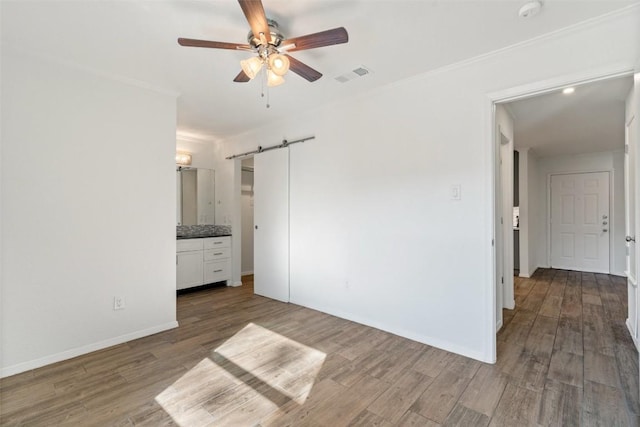 unfurnished bedroom featuring ensuite bathroom, ceiling fan, crown molding, wood-type flooring, and a barn door