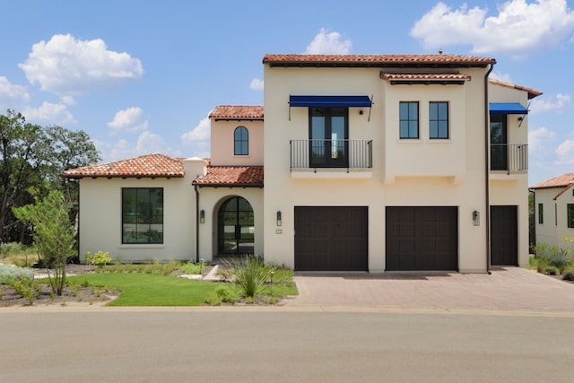 mediterranean / spanish-style home featuring a balcony, a tiled roof, an attached garage, decorative driveway, and stucco siding