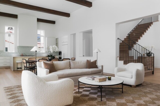 living area featuring light wood-style flooring, a towering ceiling, baseboards, stairs, and beam ceiling