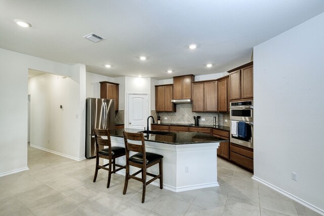 kitchen with a kitchen island with sink, a kitchen breakfast bar, decorative backsplash, dark stone countertops, and appliances with stainless steel finishes