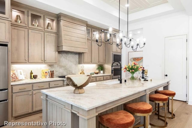 kitchen with a raised ceiling, a kitchen island with sink, a breakfast bar, and premium range hood