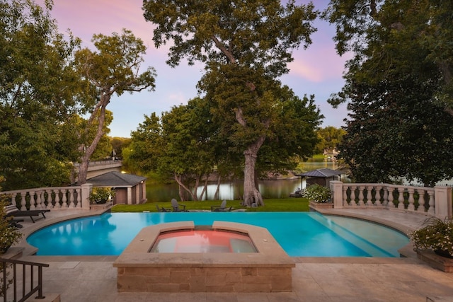 pool at dusk with a water view, a yard, and an in ground hot tub