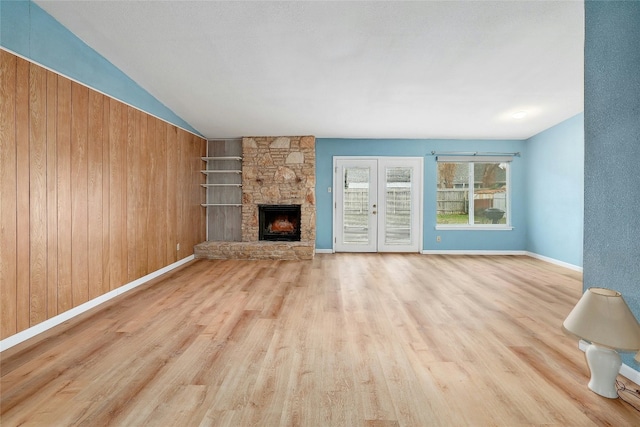 unfurnished living room with french doors, a stone fireplace, wood walls, light hardwood / wood-style floors, and vaulted ceiling