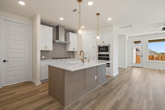 kitchen featuring stainless steel appliances, wall chimney range hood, light hardwood / wood-style floors, a kitchen island with sink, and white cabinets