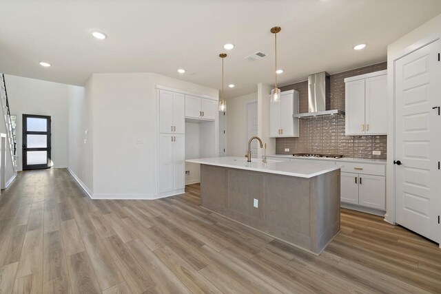 kitchen featuring wall chimney range hood, light hardwood / wood-style flooring, white cabinets, hanging light fixtures, and an island with sink