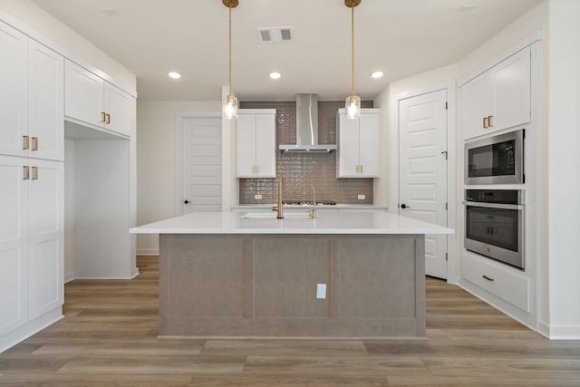 kitchen featuring white cabinets, appliances with stainless steel finishes, wall chimney exhaust hood, and an island with sink