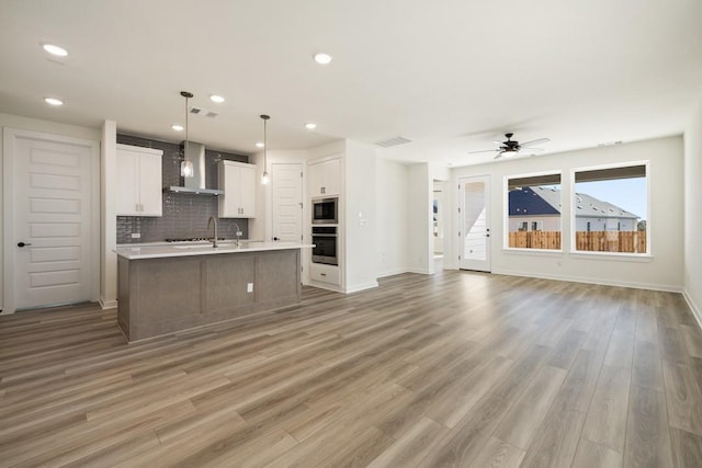 kitchen with hanging light fixtures, light hardwood / wood-style flooring, wall chimney exhaust hood, an island with sink, and white cabinetry