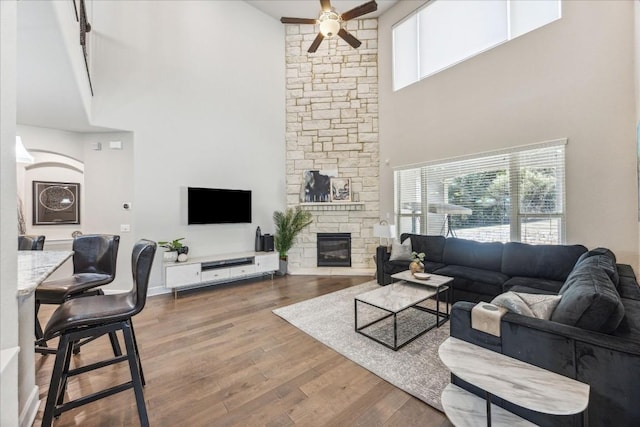 living room featuring ceiling fan, a stone fireplace, wood-type flooring, and a high ceiling