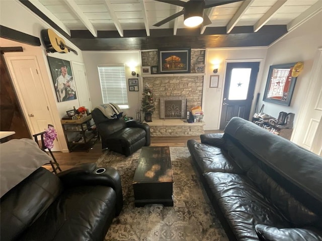 living room with beamed ceiling, wood-type flooring, a stone fireplace, and ceiling fan