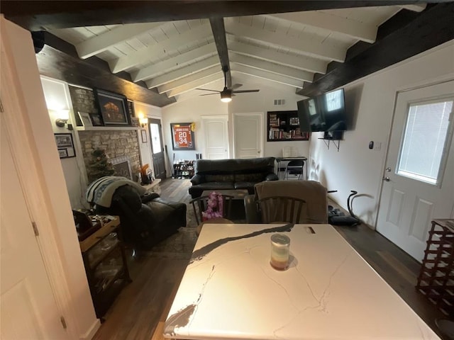 dining area with lofted ceiling with beams, ceiling fan, a stone fireplace, and dark wood-type flooring