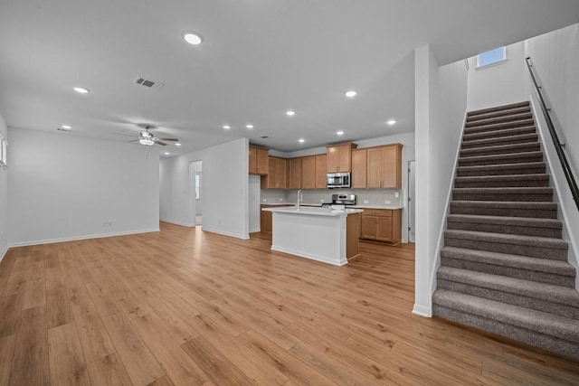 kitchen with backsplash, stainless steel appliances, ceiling fan, a center island with sink, and light hardwood / wood-style flooring