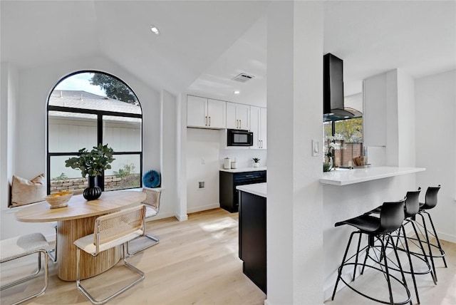 kitchen featuring wall chimney exhaust hood, kitchen peninsula, light hardwood / wood-style floors, a kitchen bar, and white cabinets