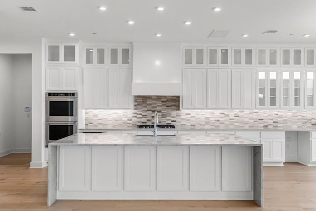 kitchen with a large island with sink, double oven, light stone countertops, decorative backsplash, and white cabinets