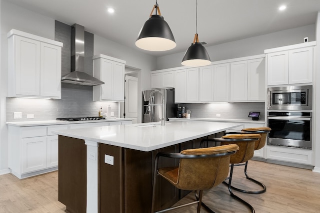 kitchen with pendant lighting, wall chimney exhaust hood, an island with sink, light hardwood / wood-style floors, and stainless steel appliances