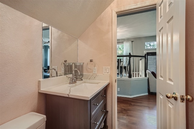 half bath with vanity, wood finished floors, baseboards, toilet, and a textured wall