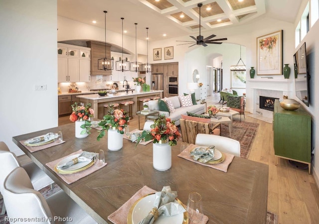 dining space with coffered ceiling, a premium fireplace, light hardwood / wood-style flooring, beamed ceiling, and ceiling fan with notable chandelier