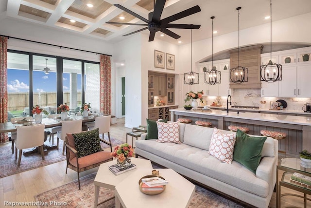 living room featuring beam ceiling, coffered ceiling, light hardwood / wood-style flooring, a towering ceiling, and ceiling fan with notable chandelier