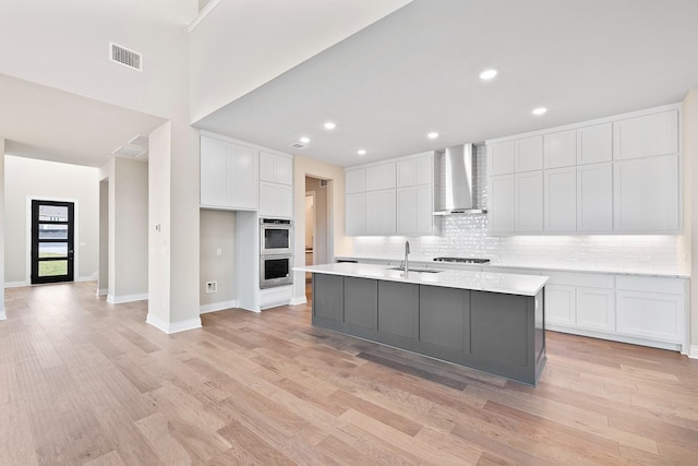 kitchen featuring sink, wall chimney exhaust hood, double oven, a kitchen island with sink, and white cabinets