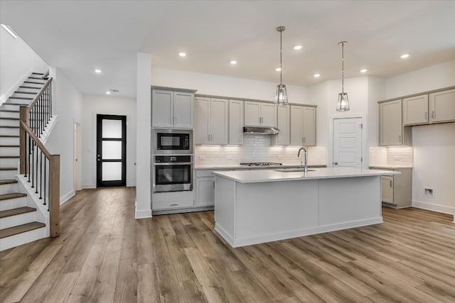 kitchen with appliances with stainless steel finishes, gray cabinetry, a center island with sink, decorative light fixtures, and light wood-type flooring