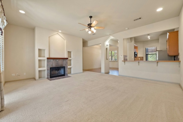 unfurnished living room with a tiled fireplace, ceiling fan, built in shelves, and light colored carpet