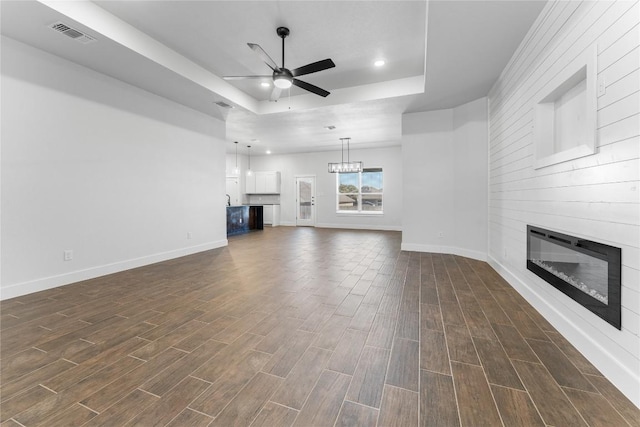 unfurnished living room featuring a raised ceiling, a fireplace, ceiling fan with notable chandelier, and dark hardwood / wood-style floors