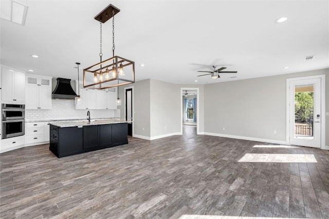 kitchen with a kitchen island with sink, custom range hood, white cabinets, decorative light fixtures, and stainless steel double oven