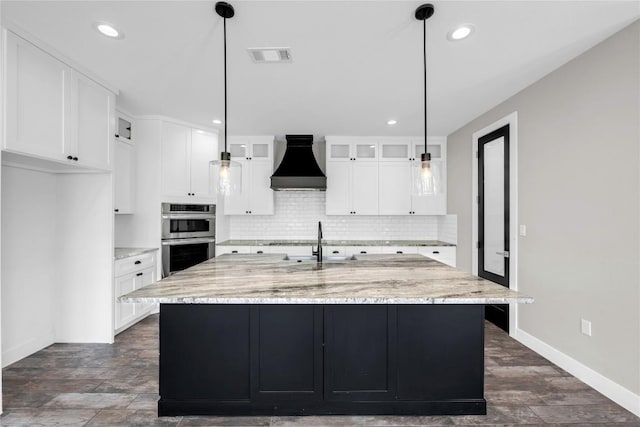 kitchen with premium range hood, double oven, an island with sink, white cabinets, and light stone counters