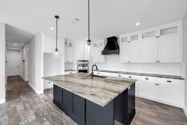 kitchen featuring white cabinetry, hanging light fixtures, custom range hood, and a center island with sink