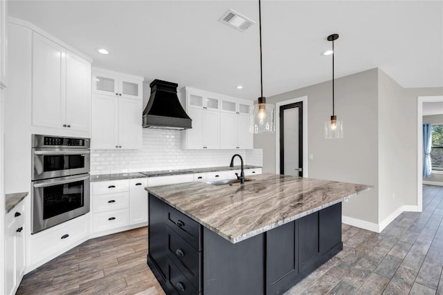 kitchen featuring pendant lighting, white cabinetry, custom range hood, a center island with sink, and stainless steel double oven