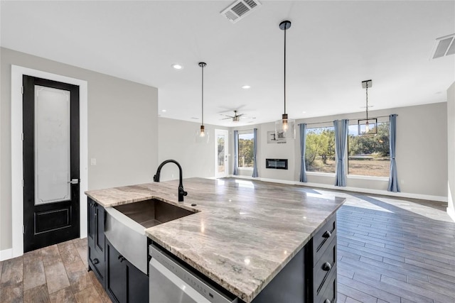 kitchen with light hardwood / wood-style flooring, a kitchen island with sink, light stone counters, decorative light fixtures, and stainless steel dishwasher