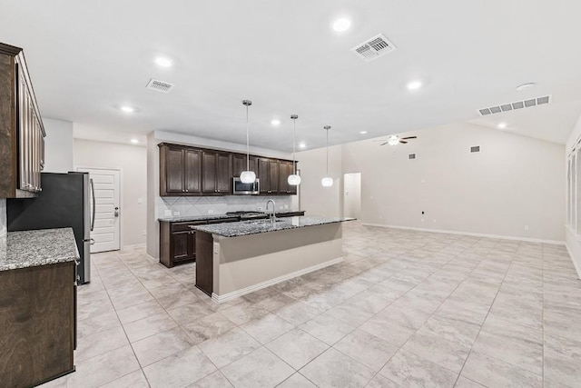 kitchen featuring appliances with stainless steel finishes, decorative light fixtures, dark brown cabinetry, light stone countertops, and a center island with sink