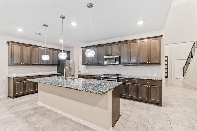 kitchen featuring appliances with stainless steel finishes, decorative light fixtures, an island with sink, dark stone countertops, and dark brown cabinets