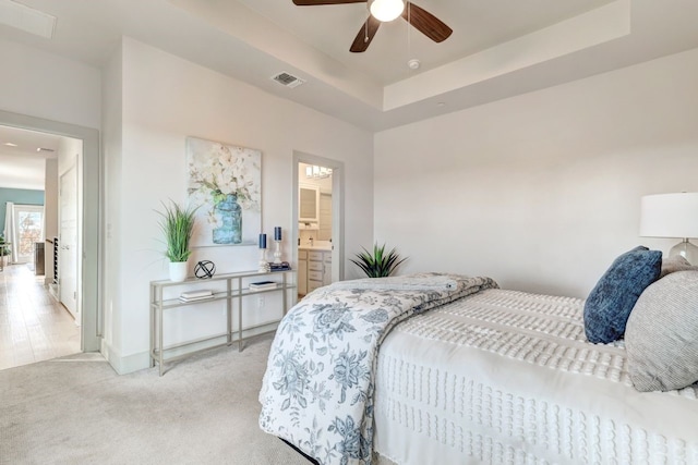 bedroom featuring a raised ceiling, ensuite bathroom, ceiling fan, and light carpet