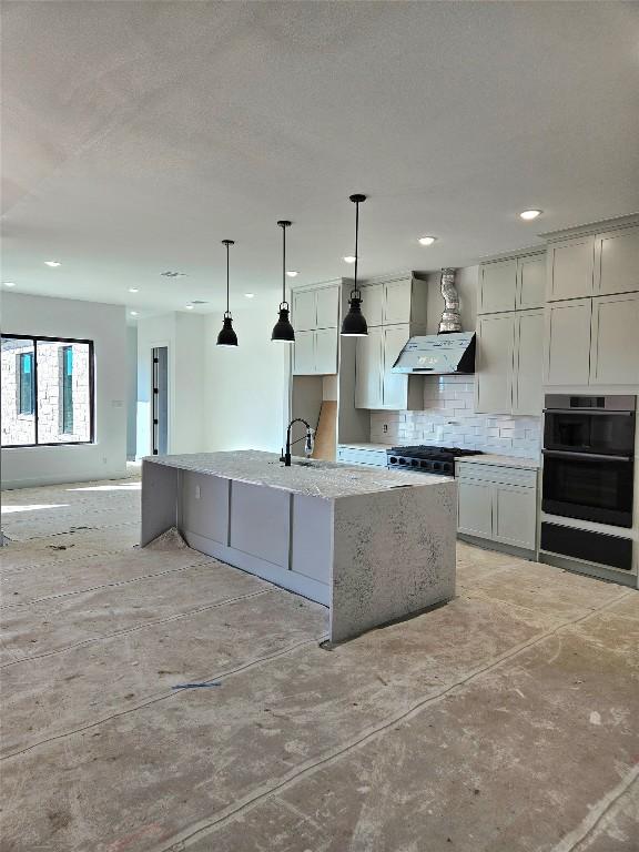 kitchen with backsplash, light stone counters, black double oven, wall chimney range hood, and an island with sink