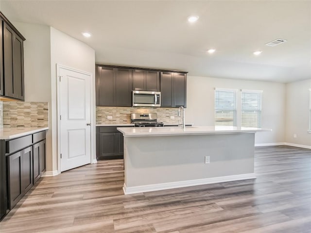 kitchen featuring backsplash, stainless steel appliances, light hardwood / wood-style flooring, and an island with sink