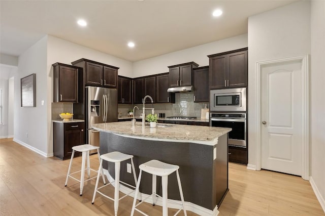 kitchen with decorative backsplash, an island with sink, appliances with stainless steel finishes, light hardwood / wood-style floors, and light stone counters