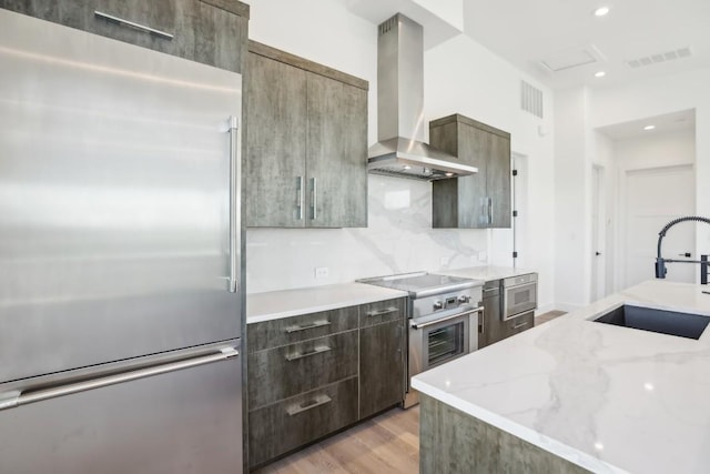 kitchen with sink, wall chimney range hood, light stone counters, light hardwood / wood-style flooring, and high end appliances