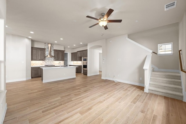 kitchen with wall chimney exhaust hood, a center island with sink, stainless steel microwave, light hardwood / wood-style floors, and backsplash