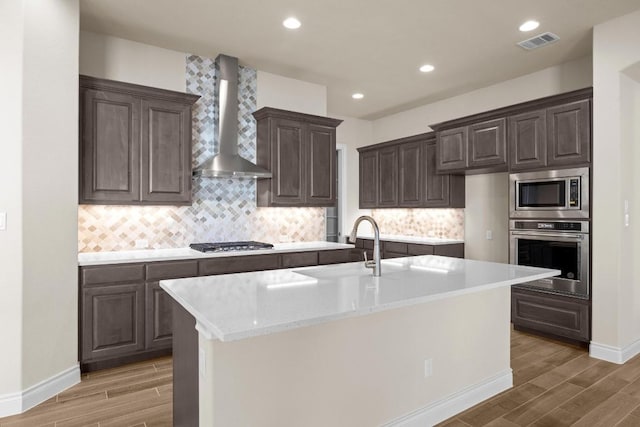 kitchen featuring hardwood / wood-style flooring, a center island with sink, wall chimney exhaust hood, and appliances with stainless steel finishes