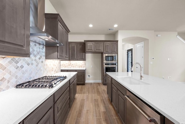 225 Skyflower Dr, Georgetown, TX 78628 - kitchen featuring wall chimney exhaust hood, stainless steel appliances, arched walkways, dark brown cabinetry, and light wood-style floors