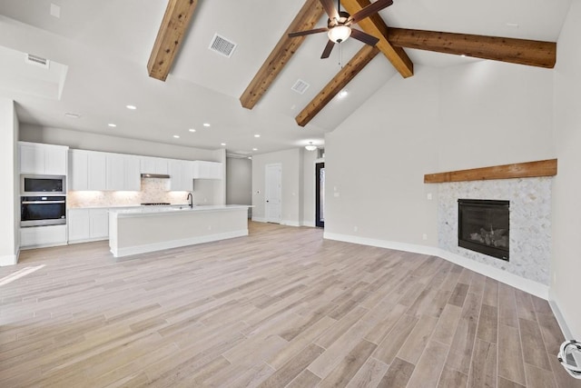 unfurnished living room with beamed ceiling, sink, a tiled fireplace, ceiling fan, and light hardwood / wood-style floors