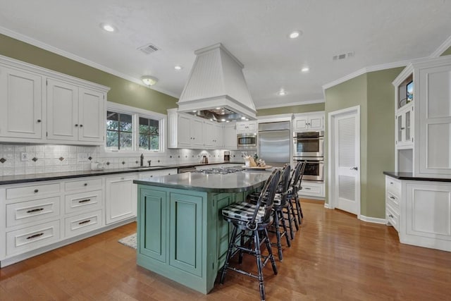 kitchen with premium range hood, white cabinets, light hardwood / wood-style flooring, built in appliances, and decorative backsplash