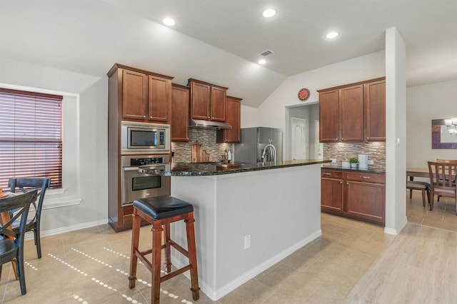 kitchen featuring a kitchen bar, dark stone counters, stainless steel appliances, vaulted ceiling, and an island with sink