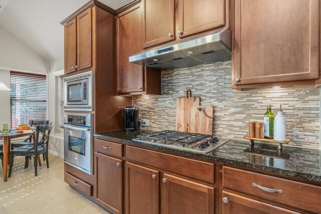 kitchen featuring lofted ceiling, backsplash, dark stone countertops, light tile patterned floors, and appliances with stainless steel finishes