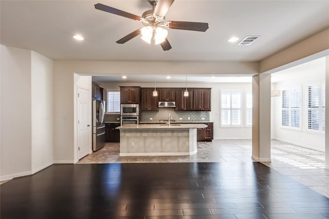 kitchen featuring tasteful backsplash, pendant lighting, wood-type flooring, appliances with stainless steel finishes, and an island with sink