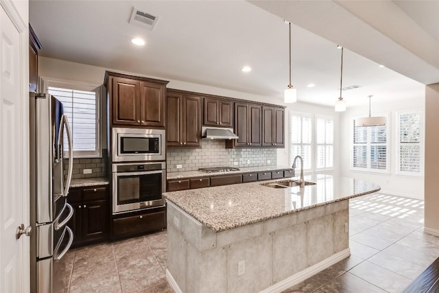 kitchen featuring decorative light fixtures, stainless steel appliances, sink, light stone counters, and a center island with sink