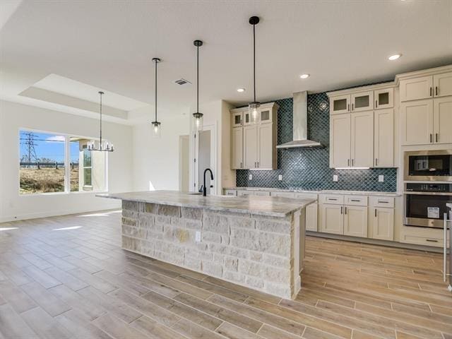 kitchen with wall chimney range hood, a raised ceiling, oven, decorative light fixtures, and a center island with sink