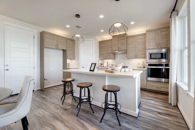 kitchen featuring hardwood / wood-style floors, backsplash, an island with sink, appliances with stainless steel finishes, and decorative light fixtures