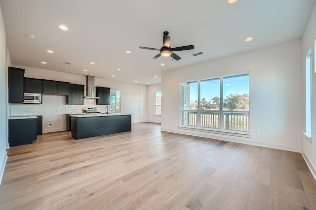 kitchen with wall chimney exhaust hood, light wood-type flooring, an island with sink, backsplash, and stainless steel microwave